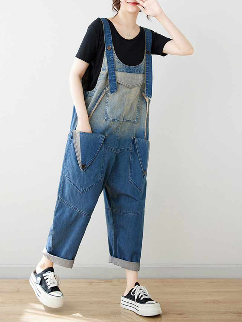 A woman wearing a black t-shirt and blue wide-leg bib's dungarees with rolled-up sleeves, standing indoors.